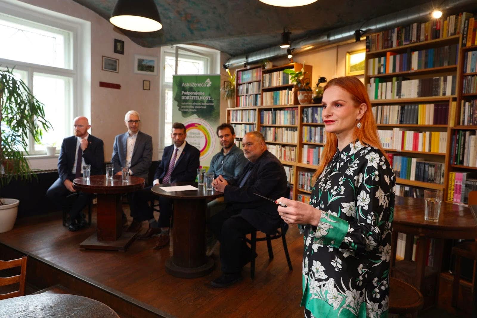 A woman speaking in a library setting with five seated men behind her near tables with glasses of water.