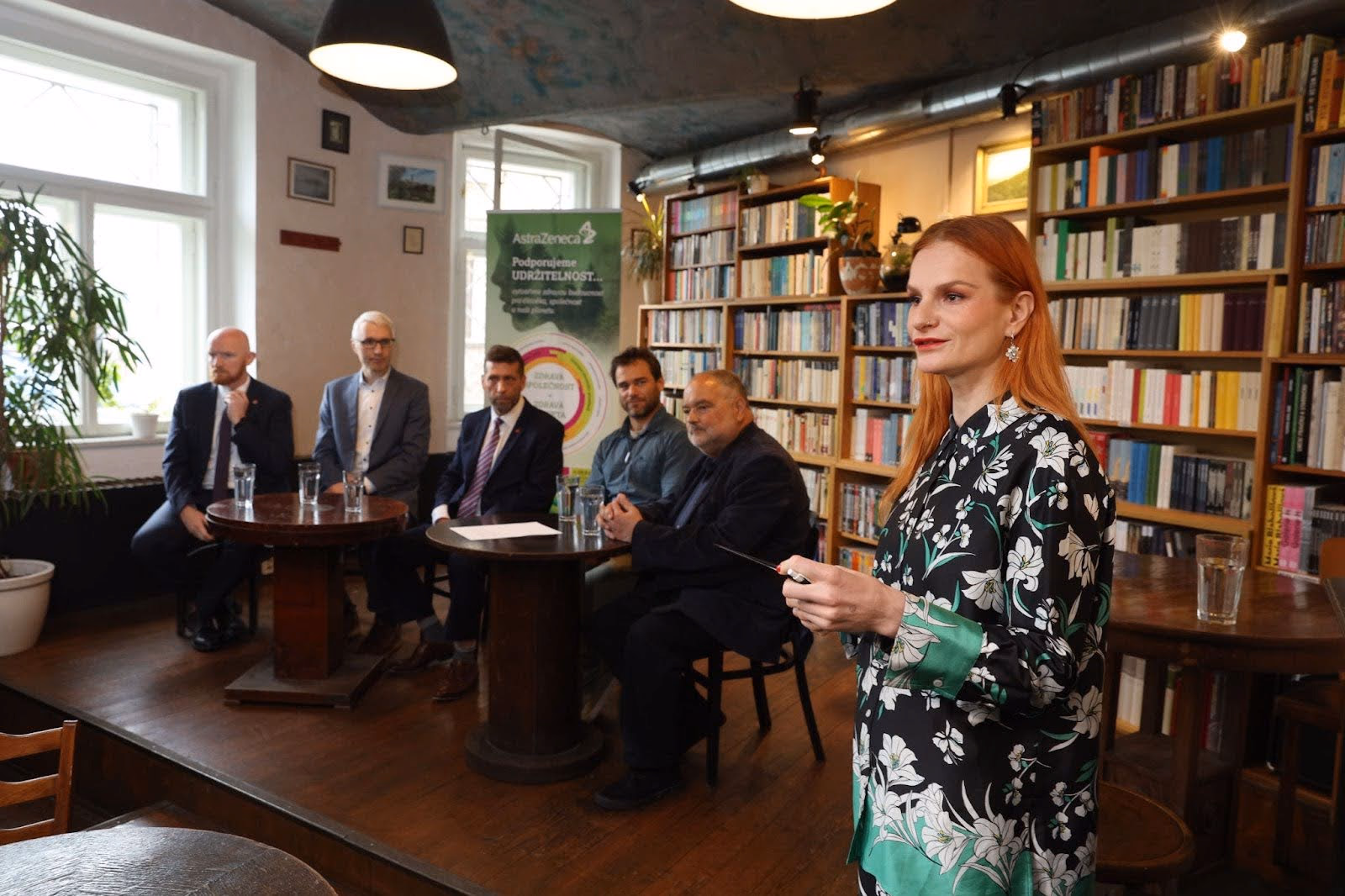 Žena hovoří v knihovně, za ní sedí pět mužů u stolů se sklenicemi vody.