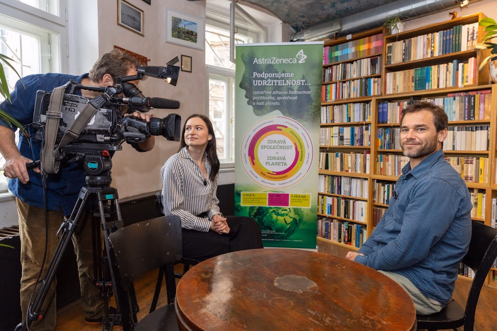 Muž s videokamerou natáčí ženu sedící u stolu s mužem v místnosti plné knihovních regálů a s plakátem AstraZeneca o udržitelnosti v pozadí.