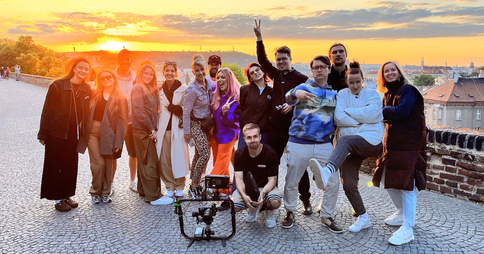 Group of smiling people posing on a cobblestone path at sunset with a camera setup in front.